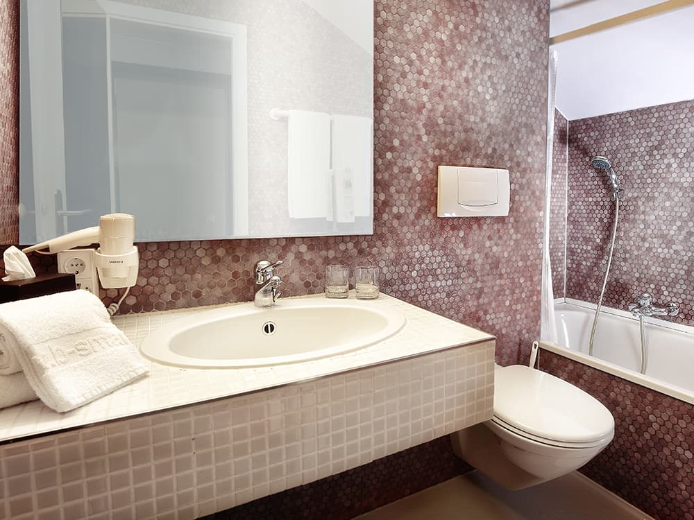 A bathroom in the b_smart motel Basel with sink, mirror, hairdryer, towels, toilet, and bathtub, decorated with hexagonal tiles