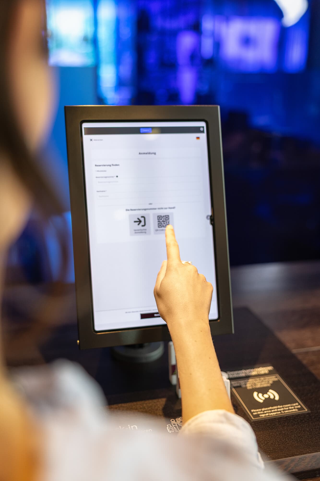 A person points at a touchscreen with a login page and QR code option at a check-in counter in Bendern.