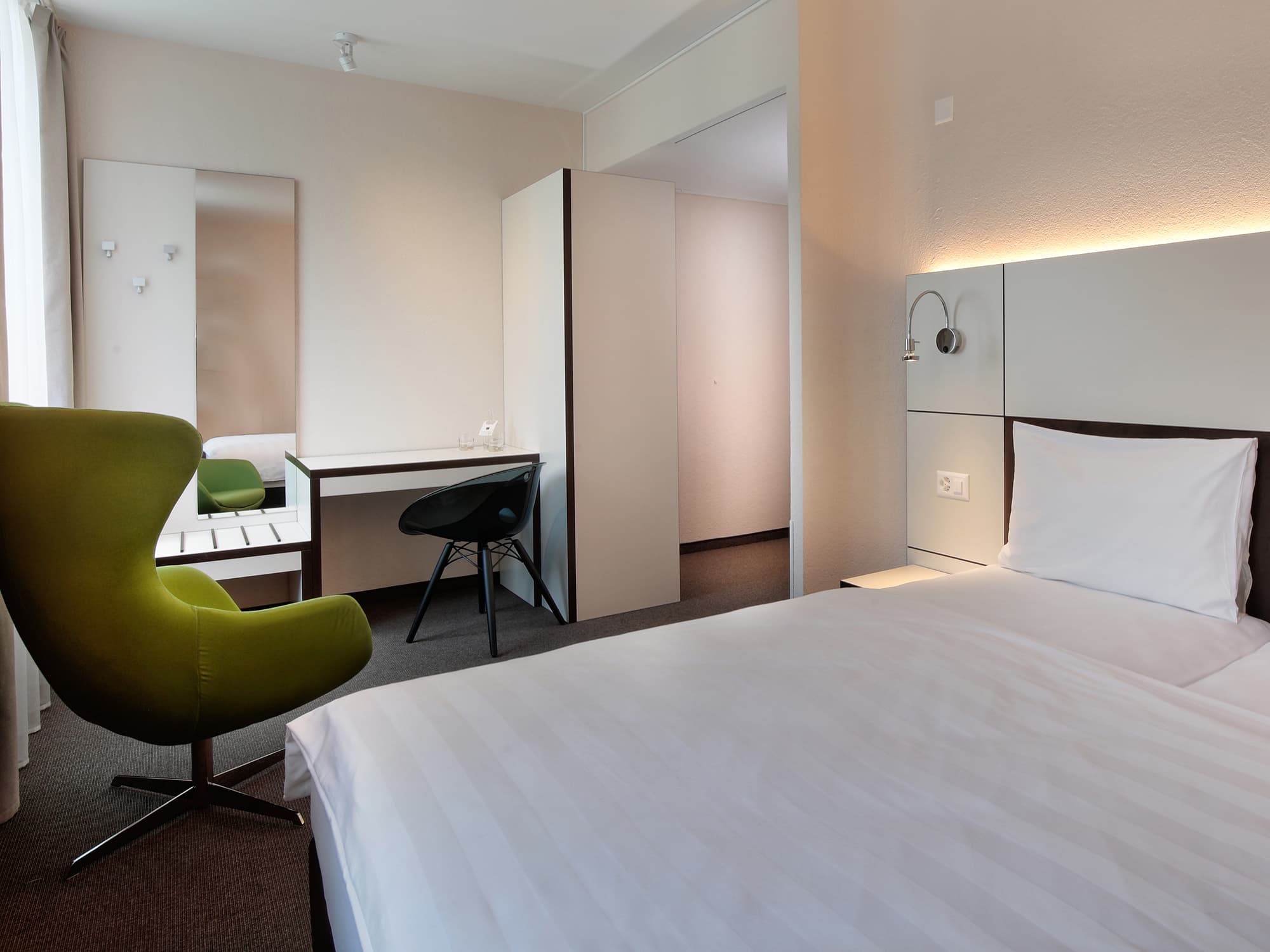 A double room in the Bsmart Motel Basel with modern design equipped with a bed a green armchair a desk a black chair and a large mirror