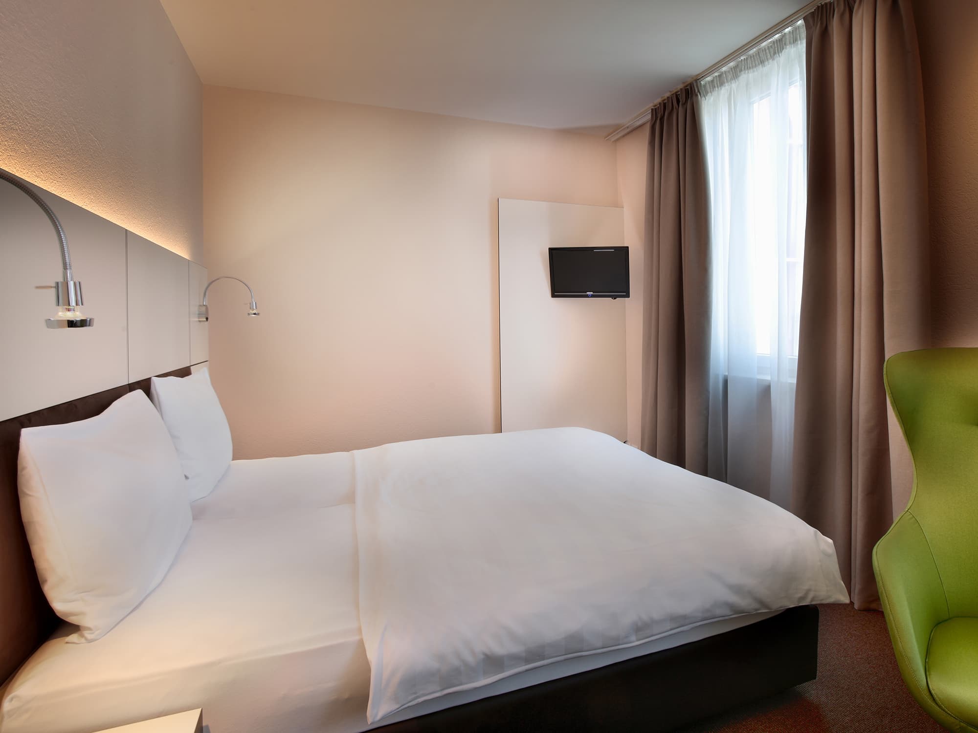 A double room in the b smart motel Basel with a large bed, soft white linens, two reading lamps, a small TV on the wall, a window with beige curtains, and a green chair in the corner.
