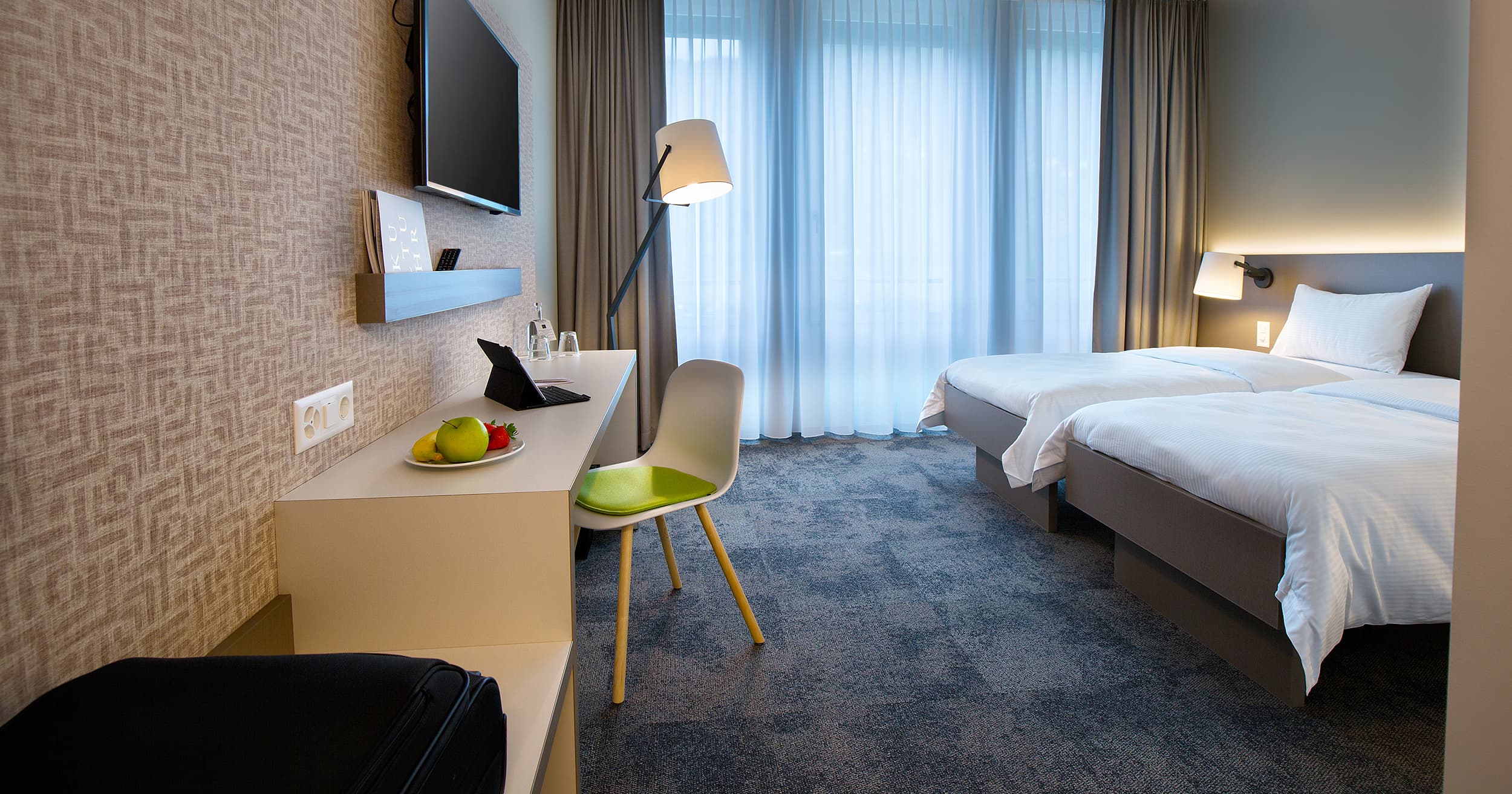 A modern double room in the B_smart Motel Landquart with two beds, a desk, a television, and a fruit bowl on a table.