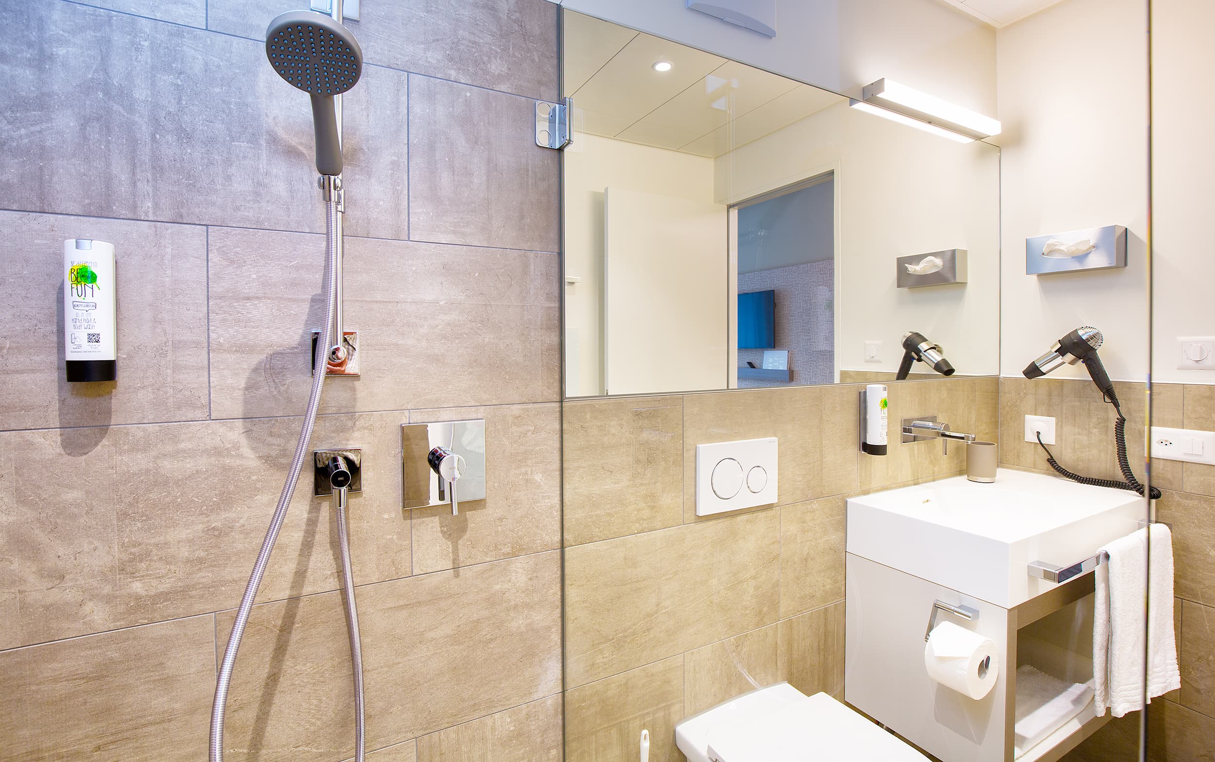 A modern bathroom in the B Smart Motel Landquart with a large shower, sink, mirror, hairdryer, and beige wall tiles.