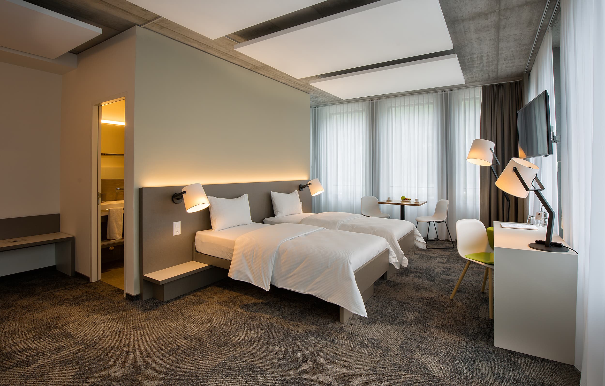 A modern hotel room at the B_smart Motel Landquart with two single beds, desk, TV, table and chairs, as well as direct access to the bathroom