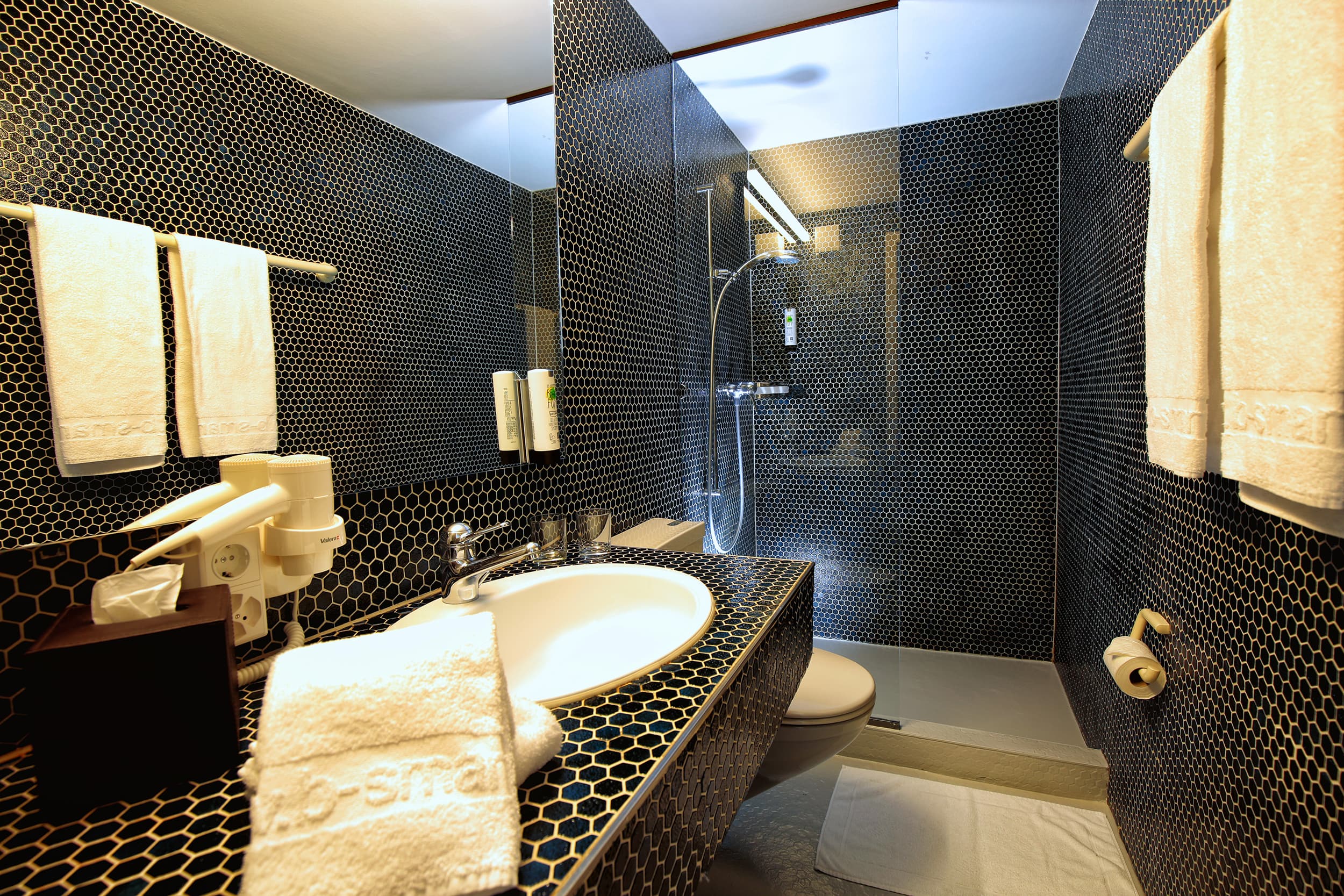 A modern hotel bathroom in the b_smart Motel Basel with a black mosaic wall, sink, shower, and white towels.