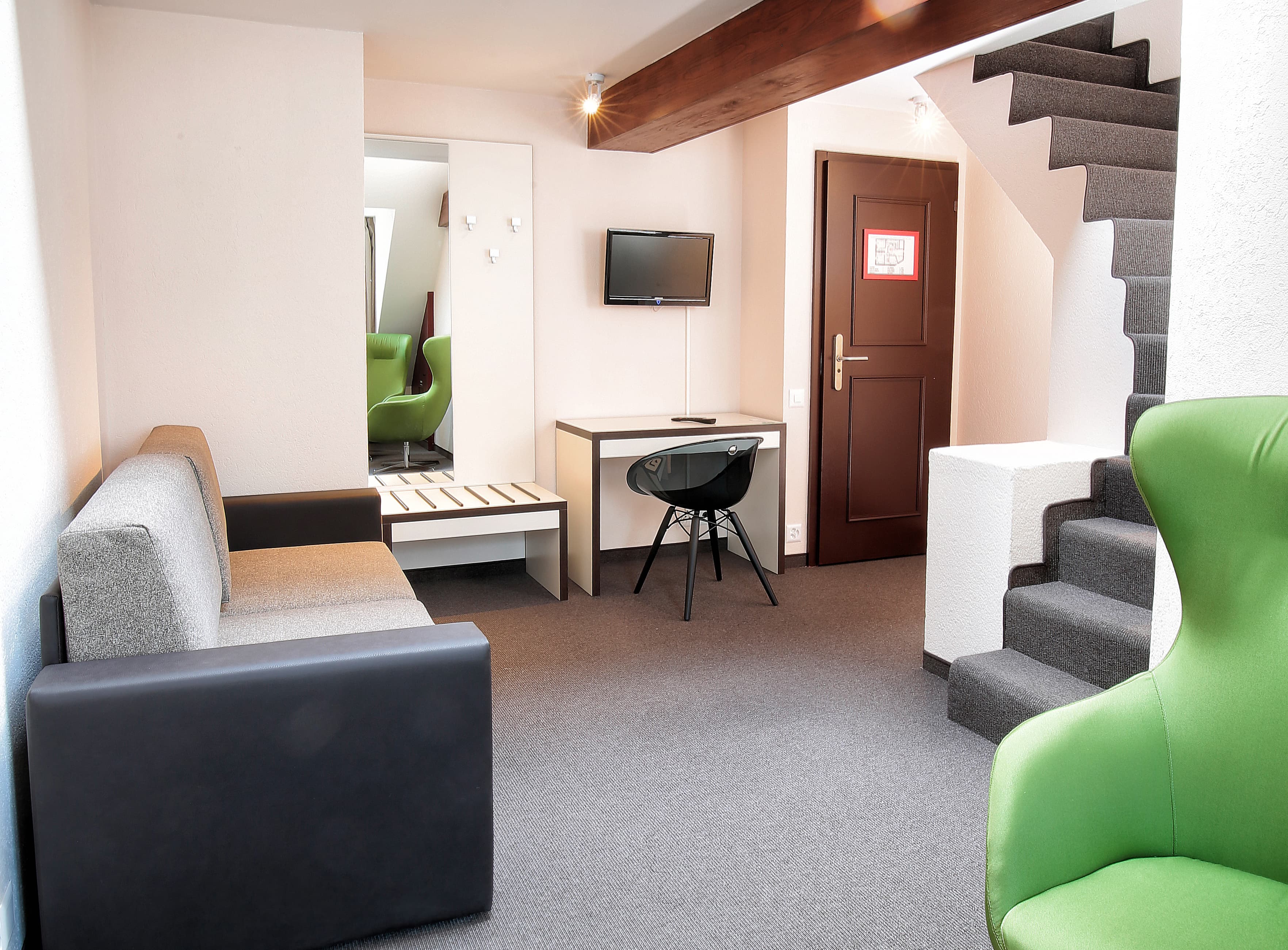 A modern room of the Junior Suite at the B Smart Motel Basel with a gray sofa, desk, television, green armchair, and staircase