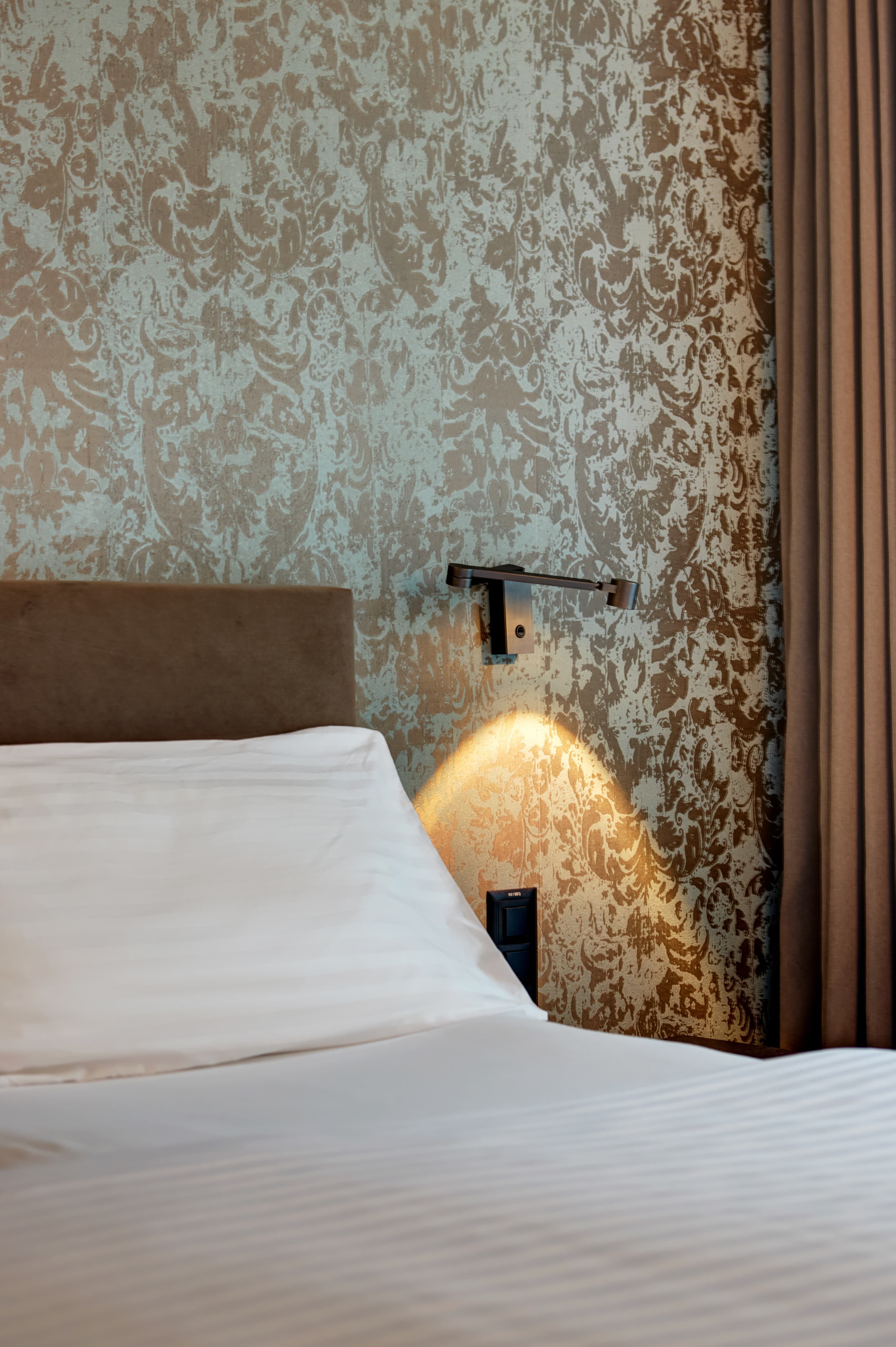 Close-up of a bed in a junior suite at the B Smart Hotel in Widnau with elegant wallpaper and a reading lamp on the wall.