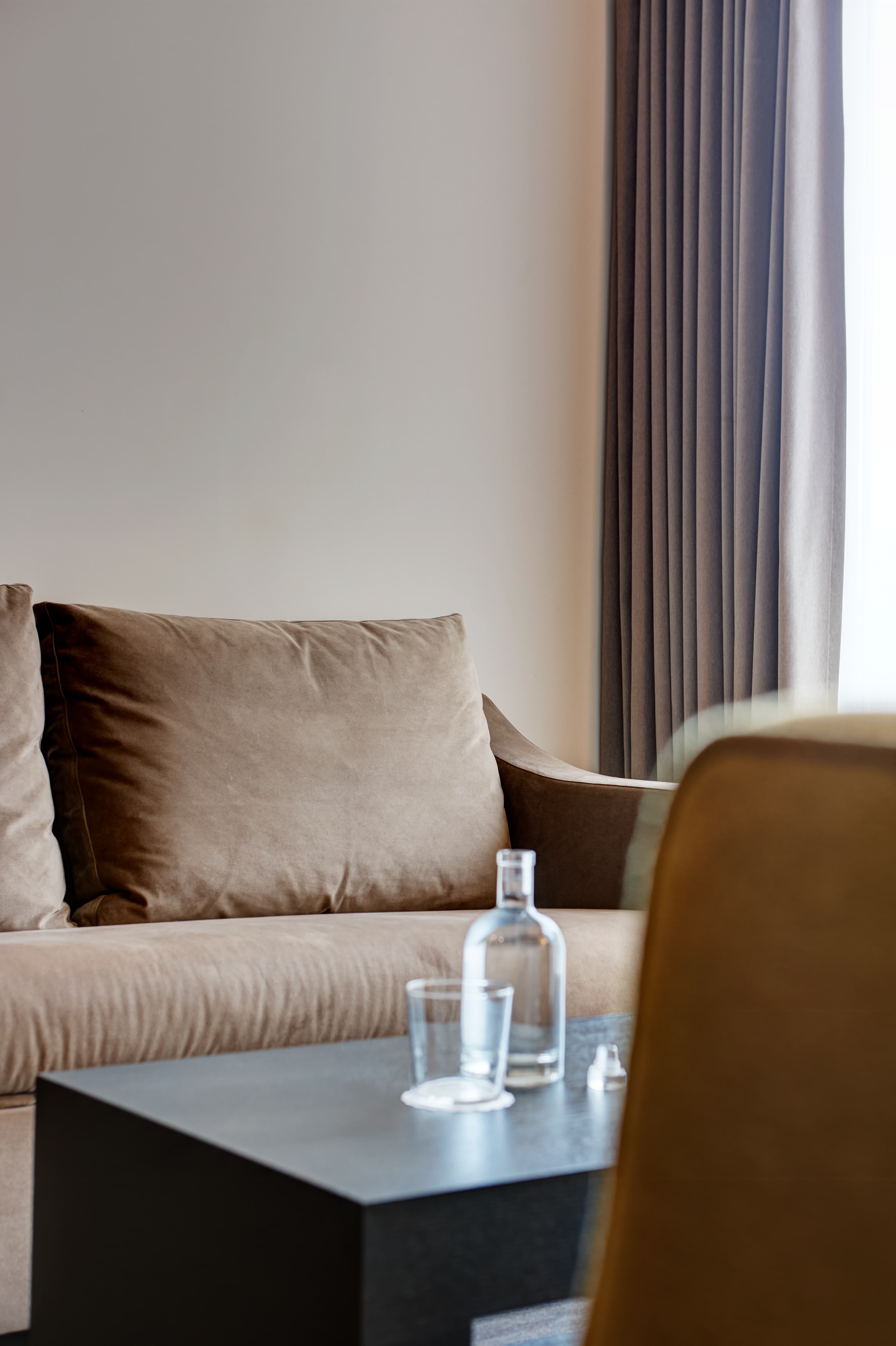 A modern hotel room in the Junior Suite of the B-smart Hotel Widnau with a brown couch, a small table, and a water bottle in front of a curtain