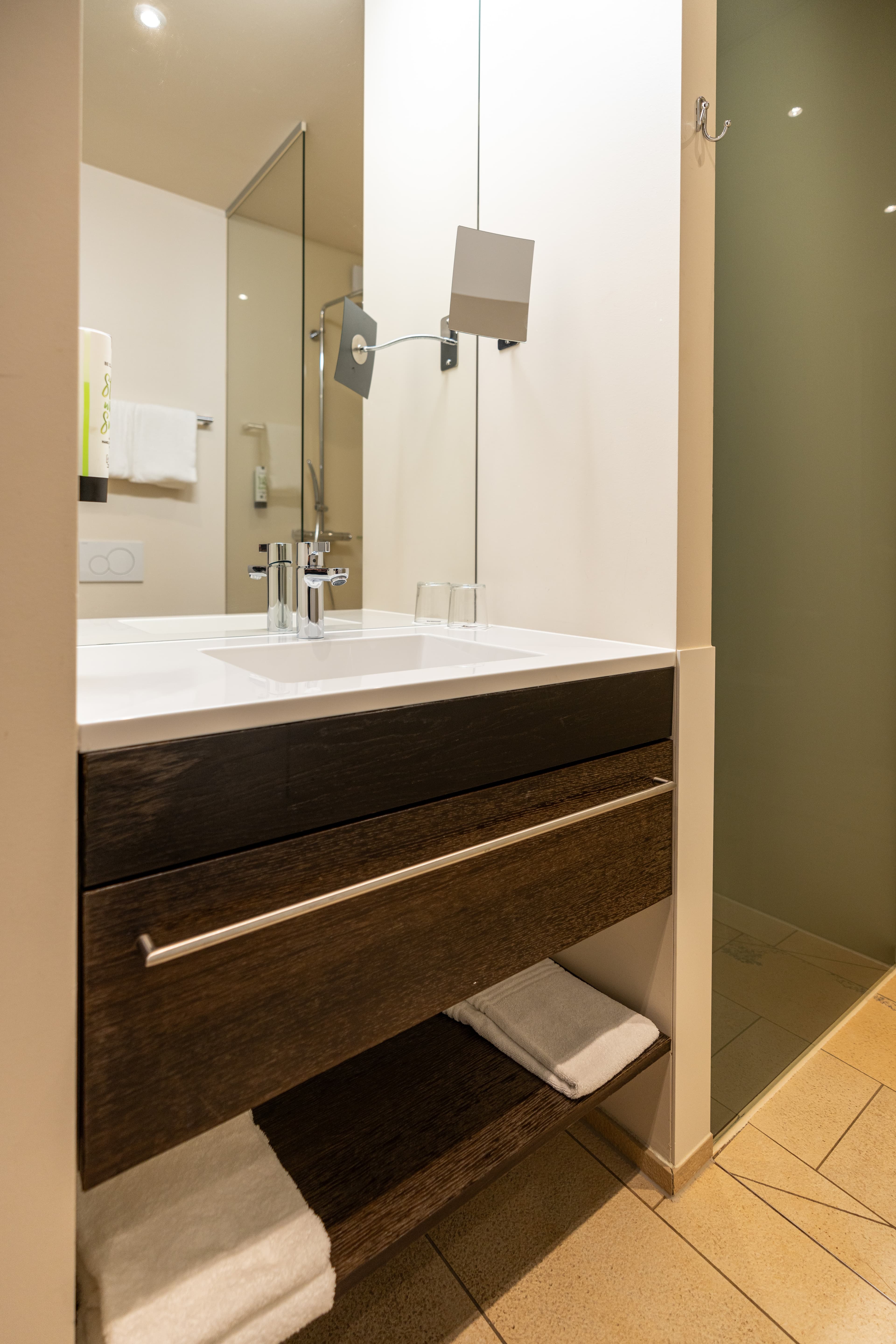 A modern bathroom in the B Smart Hotel Widnau with a sink, a mirror, a towel rack with white towels, and a shower stall