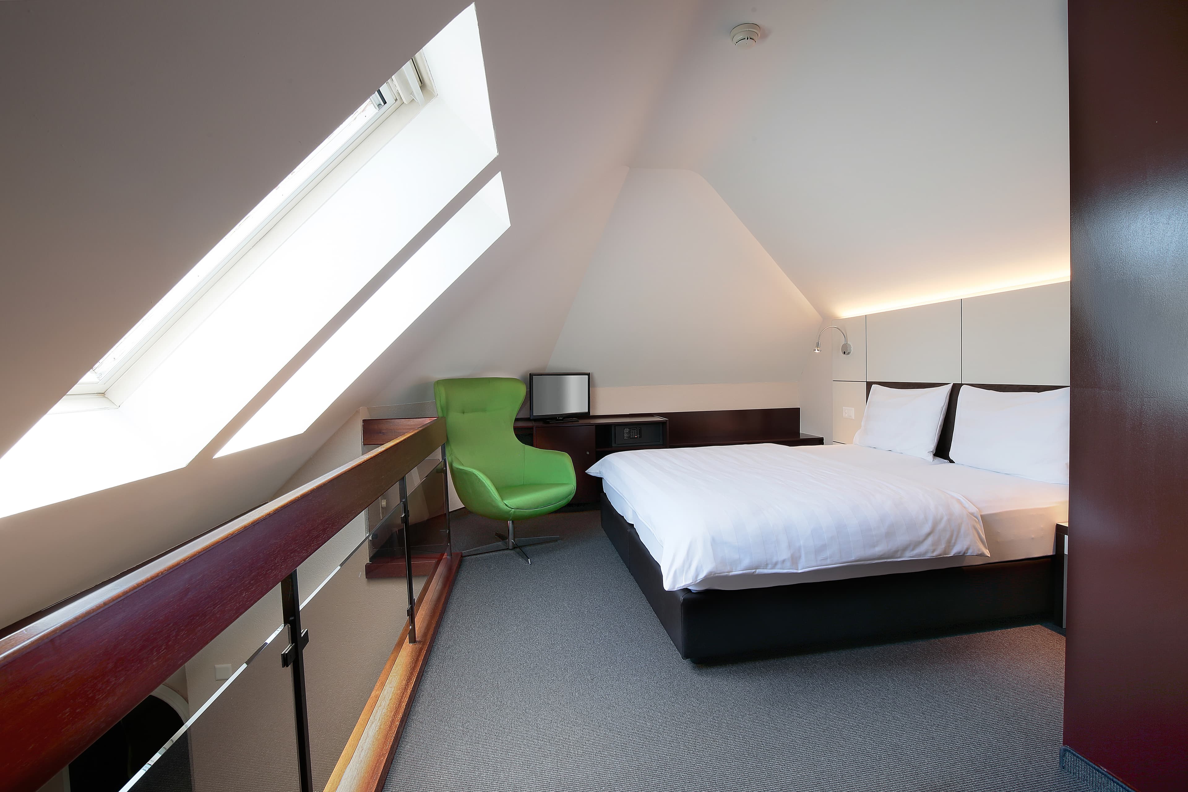 Junior Suite in the B_Smart Motel Basel with a double bed, a green chair, a small TV, and a large slanted skylight window.