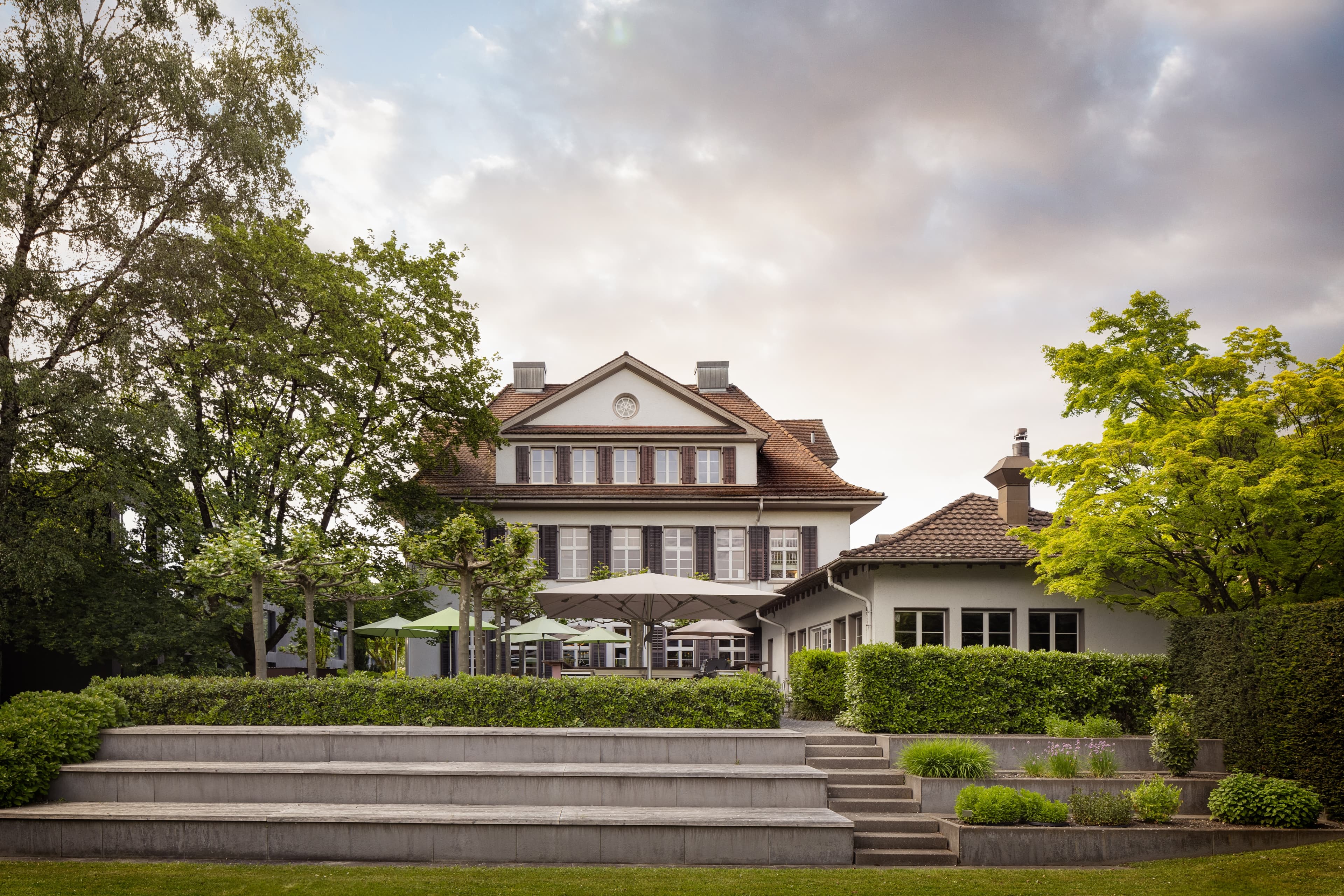 The B_Smart Hotel in Widnau with an elegant white building surrounded by green trees and plants under a cloudy sky.