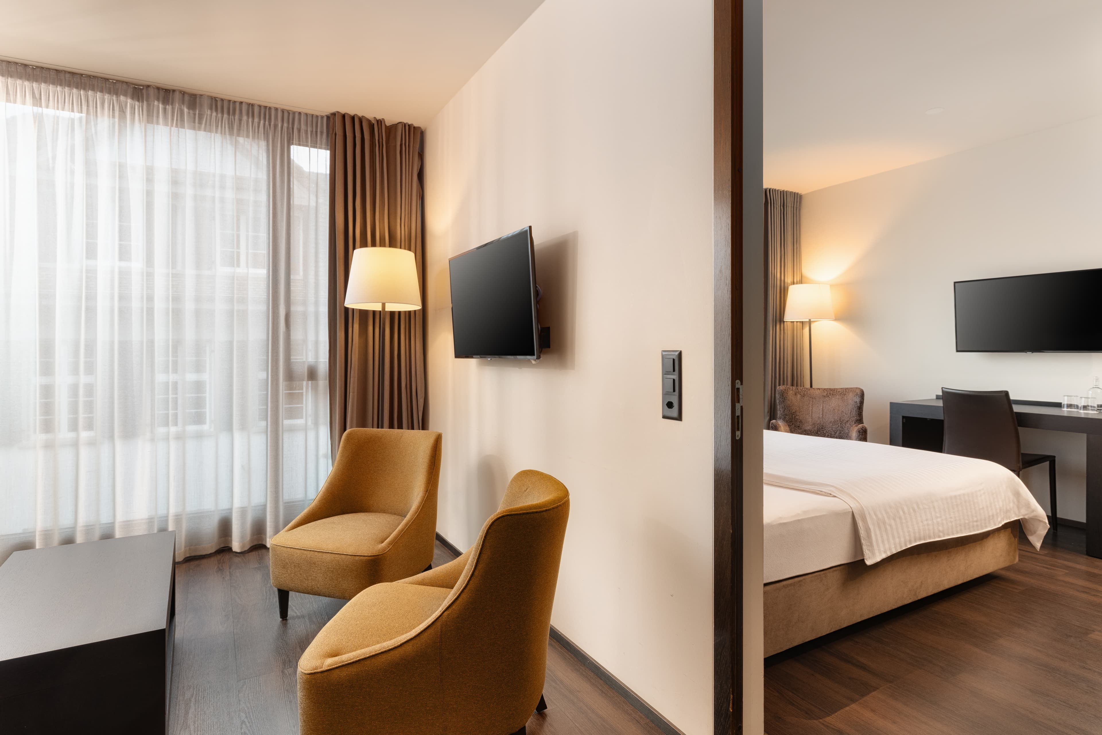 Junior Suite at the B Smart Hotels in Widnau with two armchairs, a table, a wall-mounted flat-screen TV, and an adjoining bedroom with a bed and a desk.