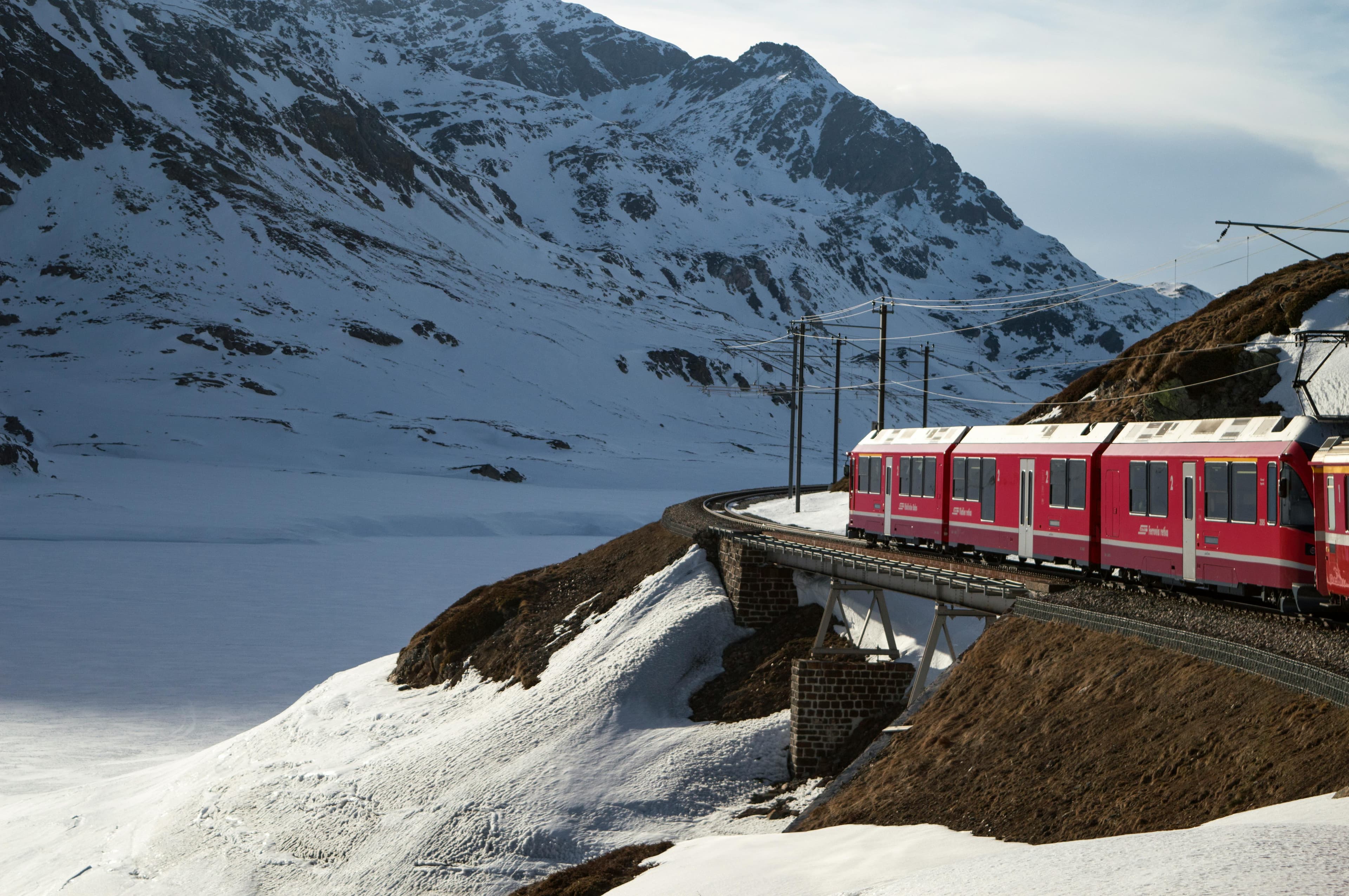 A red train of the Rhaetian Railway travels through a snow-covered mountain landscape on a viaduct in winter.
