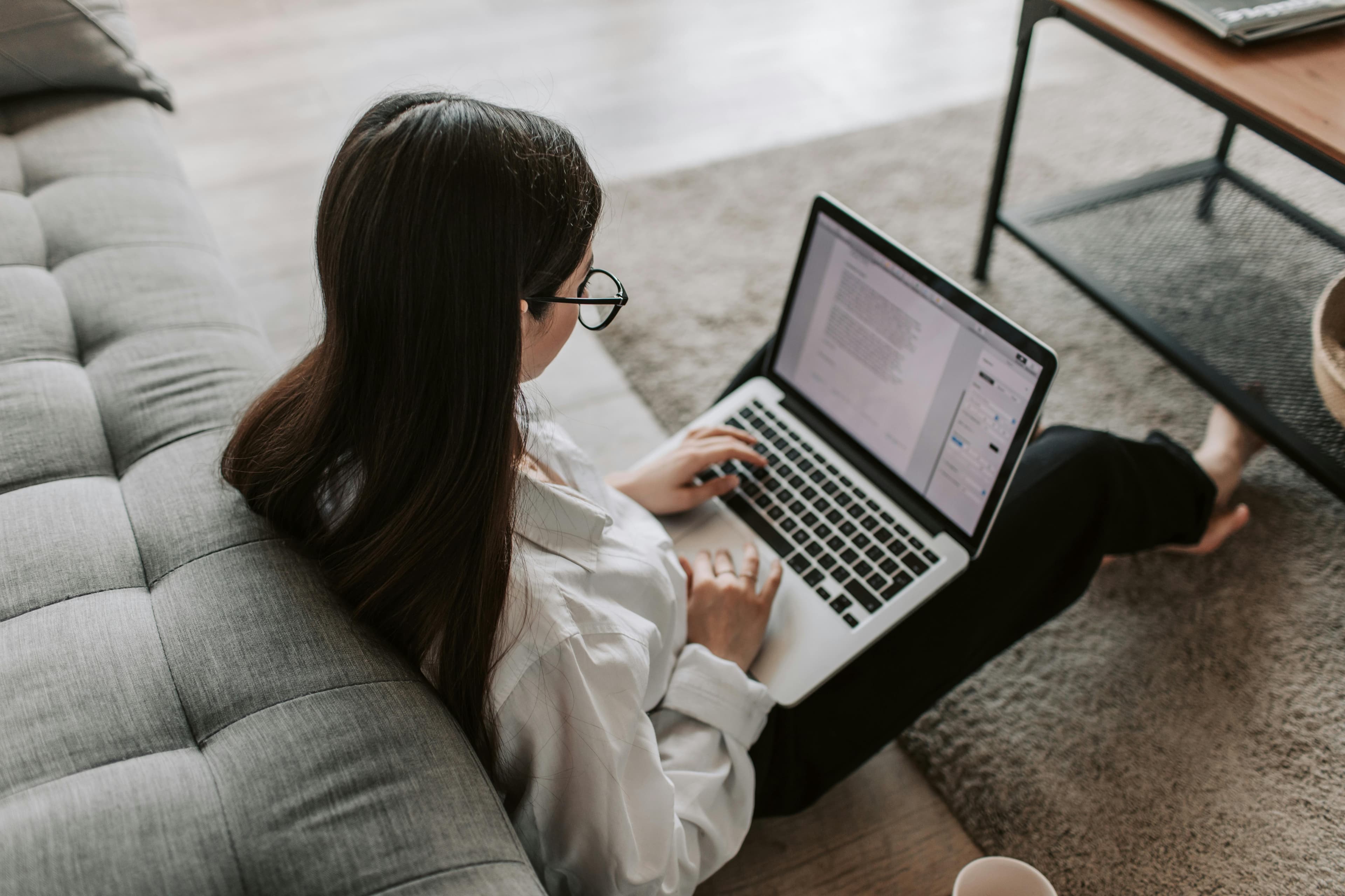 A woman sits on the floor next to a couch and works on a laptop.