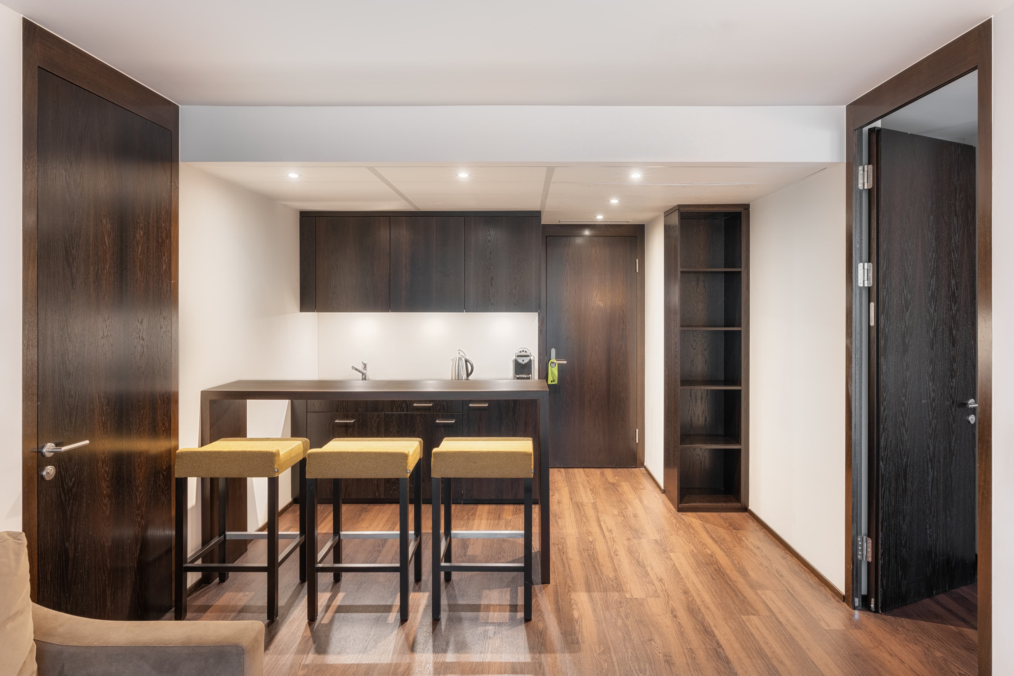 A modern hotel room with dark wood furniture, a high table, three stools, and a small kitchenette in a junior suite at the b smart Hotel Widnau.