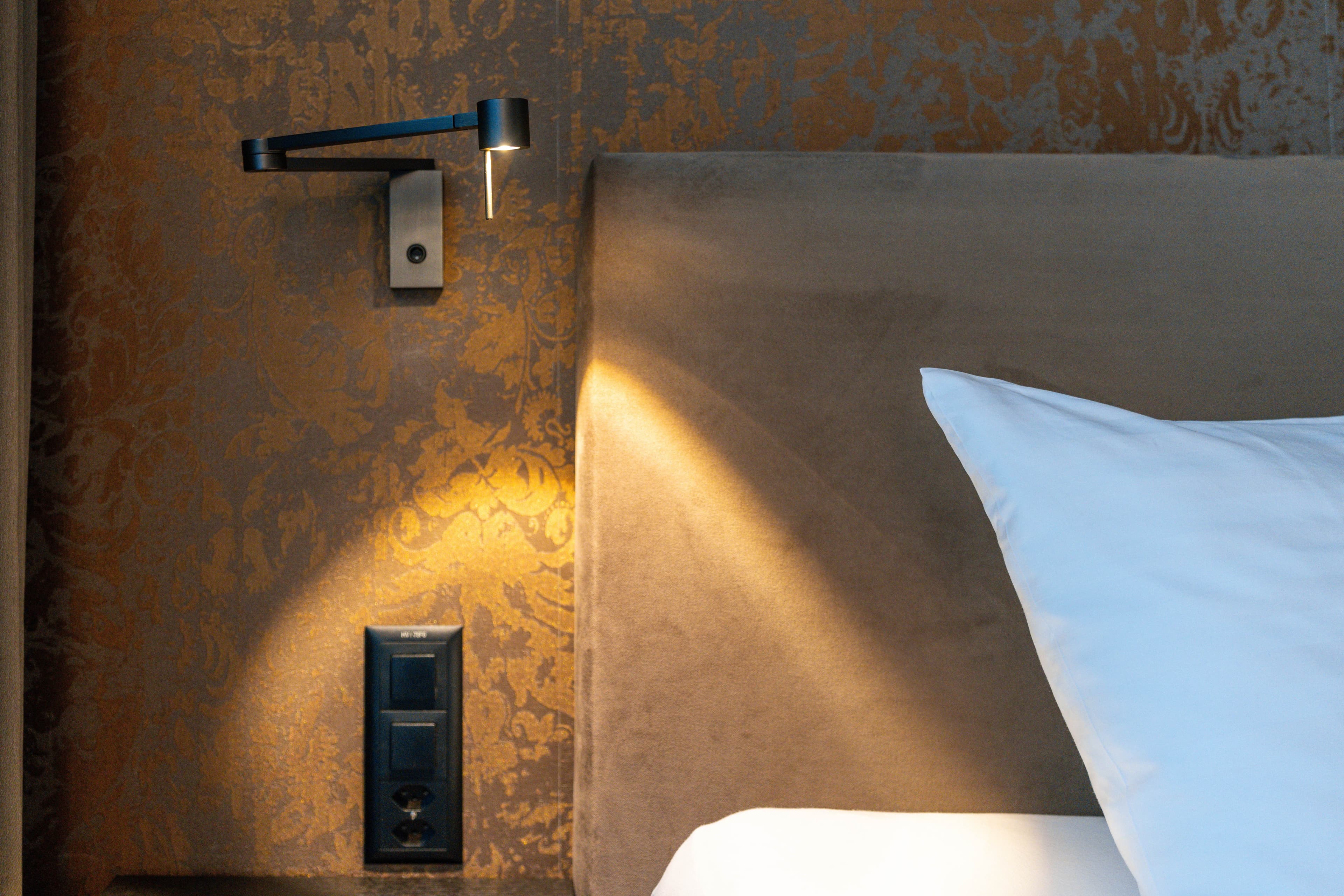 Night view of a cozy hotel room with a wall-mounted reading lamp, an outlet, and a padded headboard next to a pillow in the Bsmart Hotel Widnau.