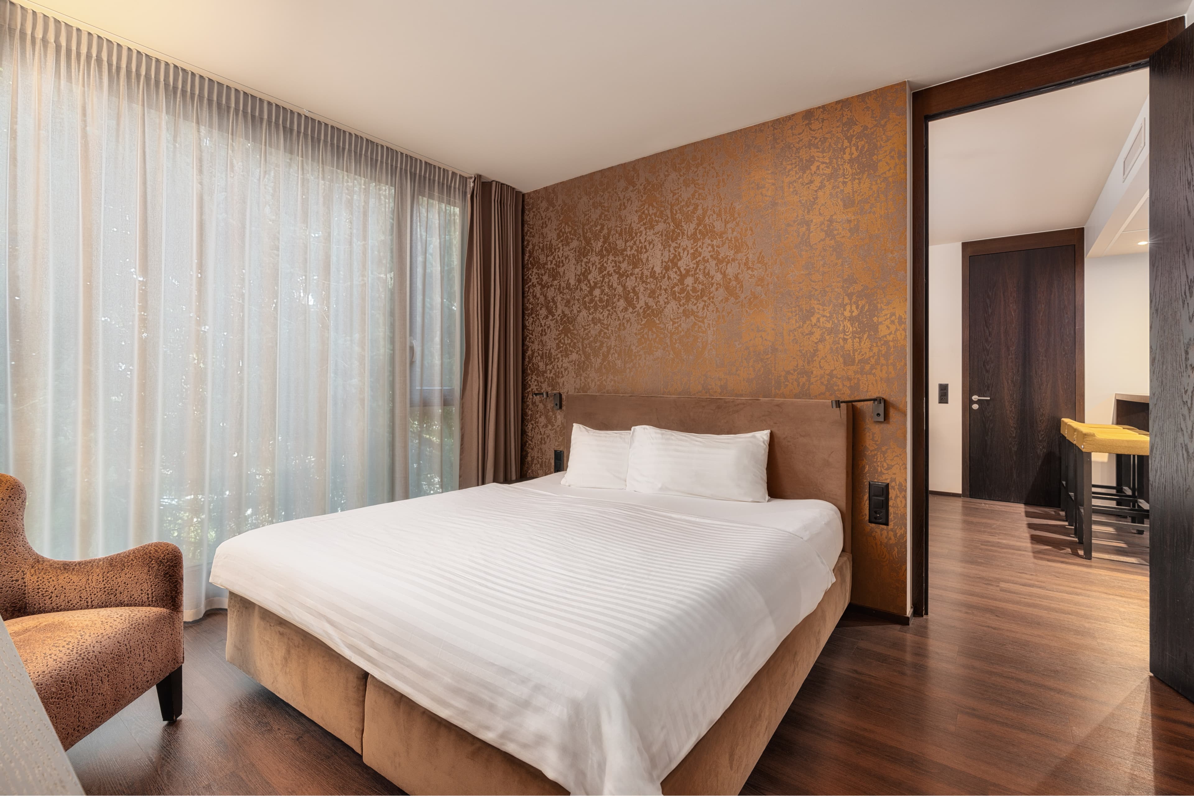 Junior suite at the b_smart Hotel Widnau with a large bed, an armchair, and a gold-colored accent on the wall.