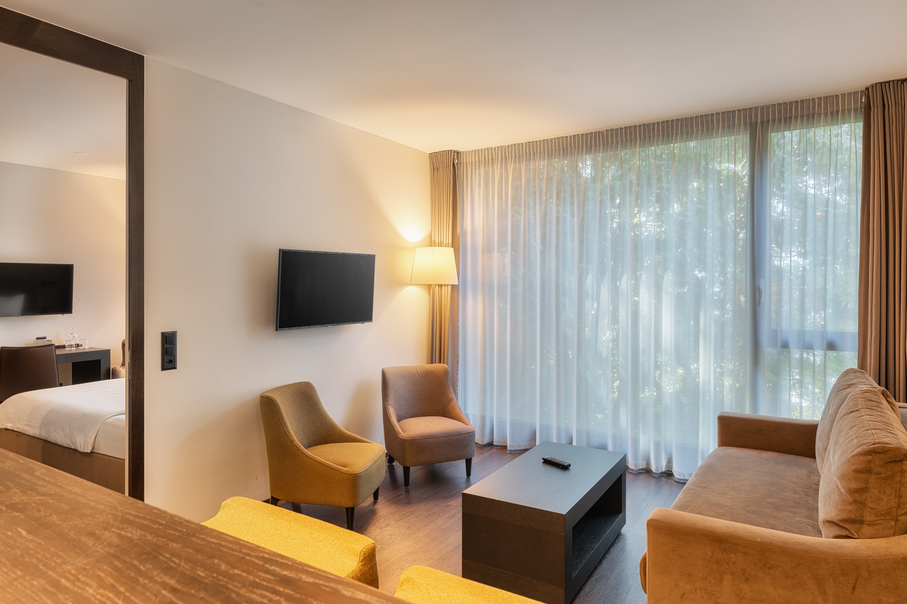 A modern hotel room in the Junior Suite at the b_smart Hotel Widnau with a sofa, armchair, coffee table, wall-mounted TV, and large windows with curtains.
