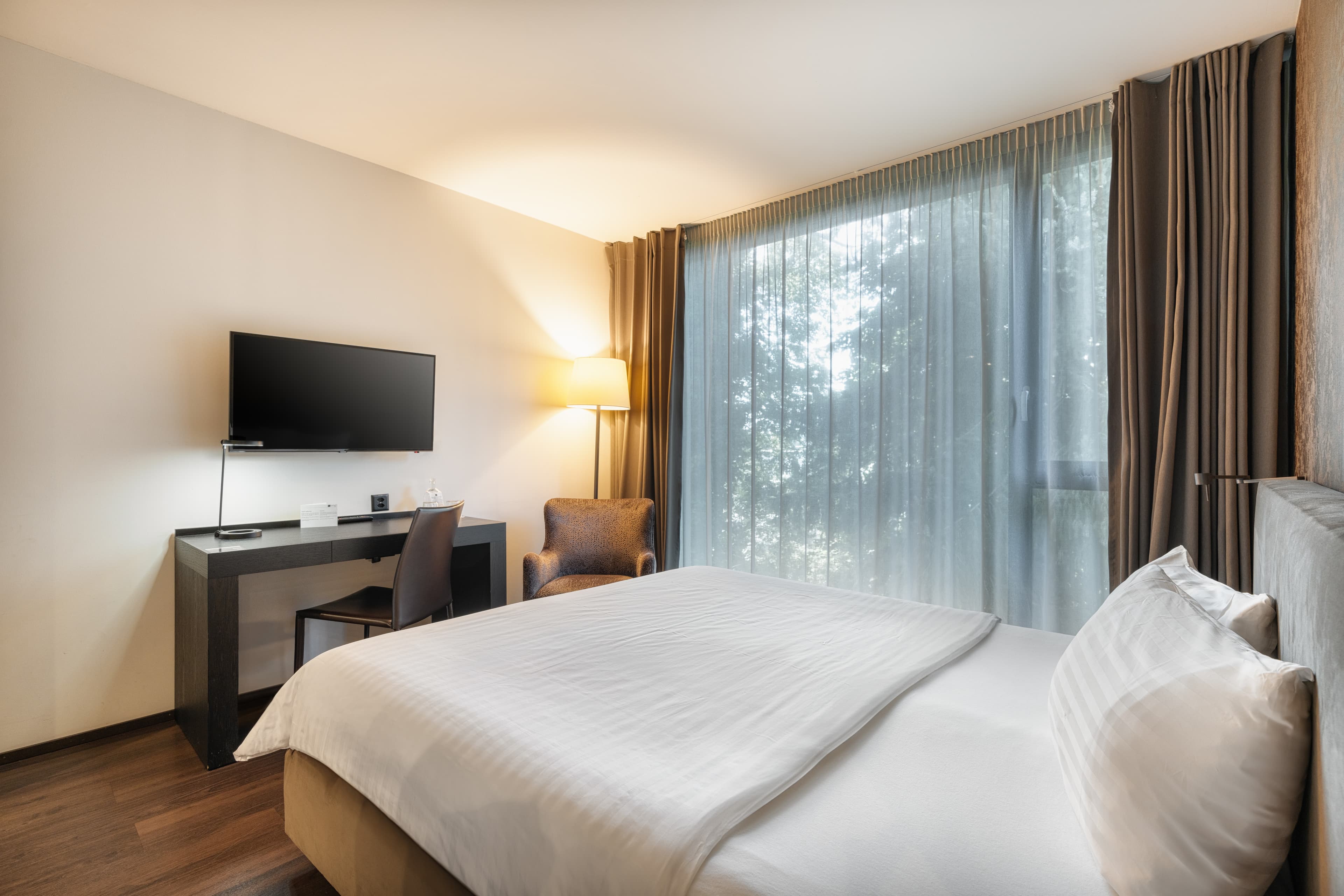 Modern double room at the B_Smart Hotel Widnau with a large bed, desk, flat-screen TV, armchair, and large windows with curtains.